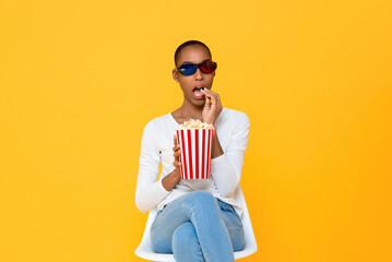 Young African American woman wearig 3D glasses enjoying watching movie and eating popcorn on isolated yellow background