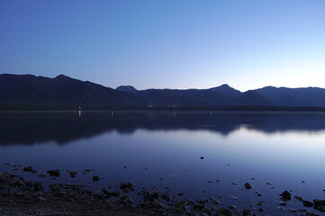 黄昏時の青い湖の風景。水面に映る空の色、遠くの山々のシルエット。
