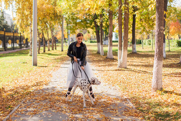 Fototapeta premium Ballerina with Dalmatian dog in the Park. Woman ballerina in a white ballet skirt and black leather jacket dancing in pointe shoes in autumn park with her spotty dalmatian dog.