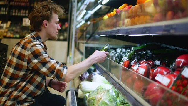A caucasian man in plaid shirt shopping for fruits and vegetables in produce department of a grocery store supermarket. Choosing perfect fresh cucumbers from the lower shelf