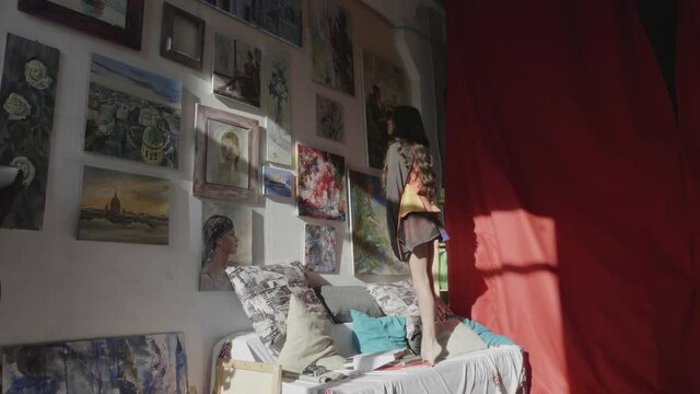 brunette girl in short dress gets on sofa to look at beautiful paintings hanging on wall in sunny art studio slow motion