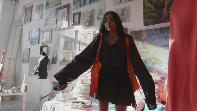 positive ballerina in orange vest and stylish dress dances near mirror in sunny art studio with own drawings slow motion