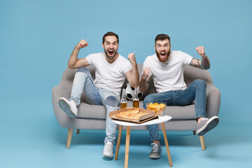 Joyful men guys friends in white t-shirt sit on couch isolated on blue background. Sport leisure...