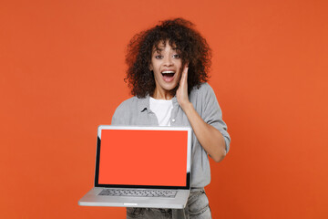 Fototapeta na wymiar Excited young african american woman in casual clothes isolated on orange background. People lifestyle concept. Mock up copy space. Hold laptop pc computer with blank empty screen, put hand on cheek.