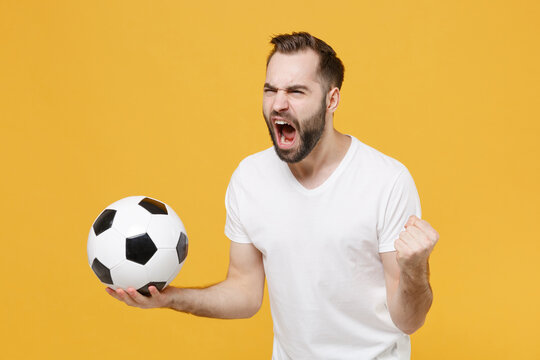 Angry Young Bearded Guy Football Fan In White T-shirt Isolated On Yellow Wall Background. People Sport Family Leisure Concept. Cheer Up Support Favorite Team With Soccer Ball Screaming Clenching Fist.