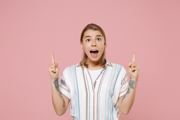 Shocked young blonde woman girl in casual striped shirt posing isolated on pastel pink background studio portrait. People emotions lifestyle concept. Mock up copy space. Pointing index fingers up.