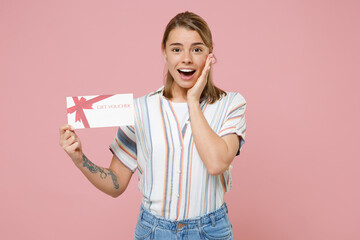 Excited young blonde woman girl in casual striped shirt posing isolated on pink wall background studio portrait. People lifestyle concept. Mock up copy space. Hold gift certificate put hand on cheek.
