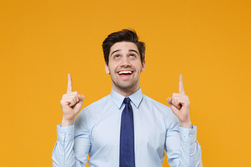 Cheerful young business man in classic blue shirt tie posing isolated on yellow background studio portrait. Achievement career wealth business concept. Mock up copy space. Pointing index fingers up.