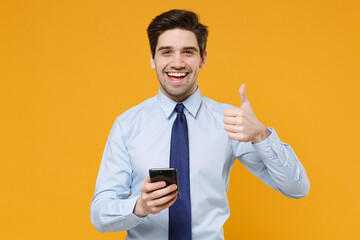 Cheerful young business man in classic blue shirt tie posing isolated on yellow background studio. Achievement career wealth business concept. Mock up copy space. Using mobile phone showing thumb up.