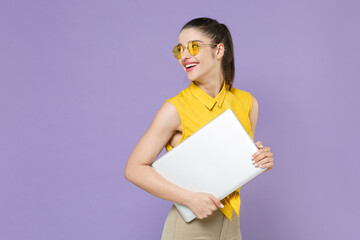 Smiling young brunette woman girl in yellow casual shirt posing isolated on violet background studio portrait. People lifestyle concept. Mock up copy space. Hold laptop pc computer, looking aside.