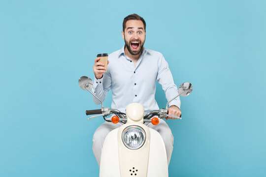 Surprised Young Bearded Man Guy In Casual Light Shirt Driving Moped Isolated On Pastel Blue Background. Driving Motorbike Transportation Concept. Mock Up Copy Space. Hold Paper Cup Of Coffee Or Tea.