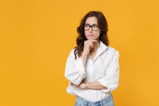 Puzzled young brunette business woman in white shirt glasses isolated on yellow background. Achievement career wealth business concept. Mock up copy space. Put hand prop up on chin, looking aside.