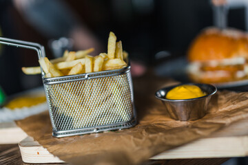 French fries in basket and cheese sauce on wooden cutting board on table. Grilled meat burger with meat on background in restaurant. Knife is stuck in the burger