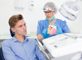 Dentist in uniform with maket of jaw is telling about healthcare in dental clinic