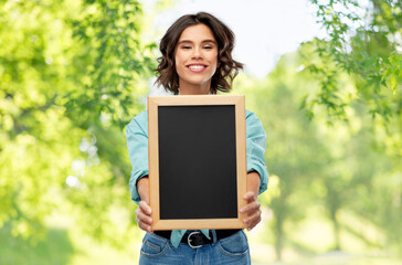 Fototapeta premium people concept - portrait of happy smiling young woman in turquoise shirt showing black chalkboard over green natural background