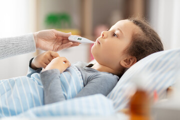family, health and people concept - mother with sick little daughter lying in bed with oral thermometer and measuring temperature at home - obrazy, fototapety, plakaty