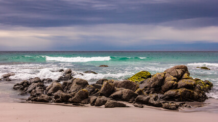 Rocks on cloudy peaceful beach, Tasmania, Australia. Light blue ocean and white sand beach with rocks and moss. Remote beach landscape.