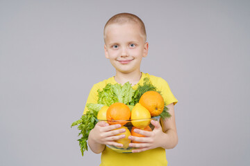 Smiling boy standing with fruits and vegetables in a plate, isolated on gray