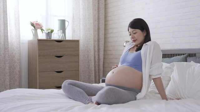asian mother-to-be sitting resting on bed is smiling with happiness while looking outside of window. pregnant woman is feeling her baby kicking and taking a look at her tummy at home. real moments