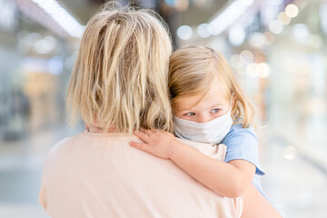 Sad baby girl wearing medical protective mask hugs mother in a public crowded place - in a shopping...