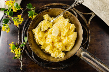 Hot corn porridge with sugar and butter on an old rustic pan and a metal vintage stand. Next to it is a sprig of flowering black currant with yellow flowers