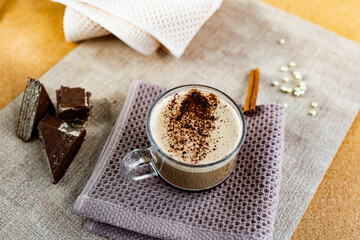 Hot cappuccino with chocolate chips on top of the foam, a cinnamon stick, Golden sugar beads and a sweet waffle next to it. Drink in a transparent Cup on a soft cloth