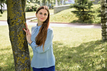 Model posing against a background of trees in sunny weather showing happiness. Photo of a caucasian brunette girl with long hair smiling in a park