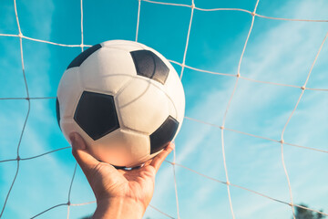 Traditional soccer ball in goal net on blue sky nature background. A hand holding classic leather football equipment to play competitive game in stadium with copy space. Sport and exercise concept