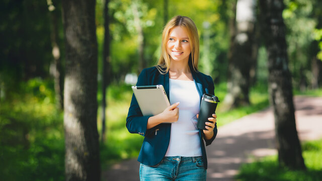 Young Happy Smiling Caucasian Adult Girl Business Student Walking Outdoor Concept In The Park Holding Reading Tablet Thinking Of Credit Finance Job In The College With Wireless Headphones Drink Coffee