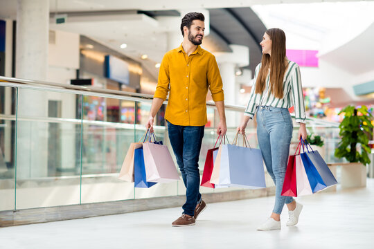 Full length photo of positive cheerful married couple man woman finish shopping in center mall go walk carry many bags packages wear striped yellow shirt denim jeans