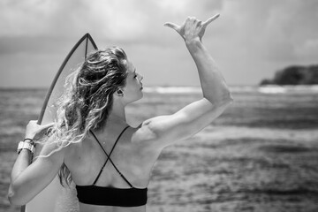 Black and white portrait of young blonde woman in bikini from her back with white surfboard showing surf sign right hand and ocean on background