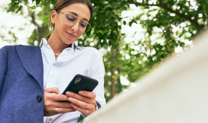 Outdoor image of young business woman wearing trendy eyewear, white shirt and blue suit, using online banking for transferring money via smartphone. Female texting email letter on mobile phone outside