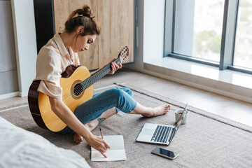 Image of young woman writing notes while playing acoustic guitar at home