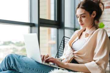 Image of focused woman working with laptop while sitting in armchair