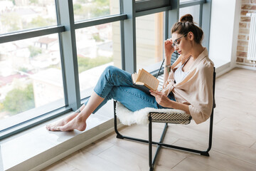 Image of focused woman reading book while sitting in armchair