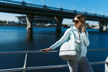A young woman with a sports bag, in a tracksuit, walking along the promenade, listening music and preparing for street sports.