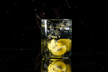 A glass of water placed on a reflective surface with black background and water is splashing after dropping a lemon in the glass. Summer concept