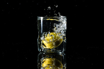 A glass of water placed on a reflective surface with black background and water is splashing after dropping a lemon in the glass. Summer concept