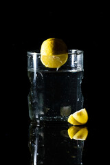 A glass of water placed on a reflective surface in a dark background with a cut lemon placed in front of it and an intact lemon on the edge of the glass