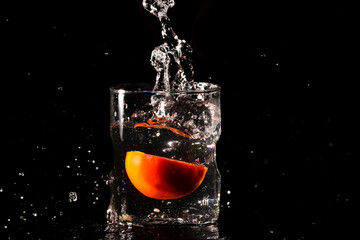 A glass of water in a dark background and water being splashed with a piece of cut tomato dropped inside the glass