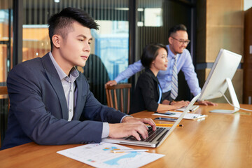 Handsome young man sitting at office table searching something on his laptop while his colleagues working together