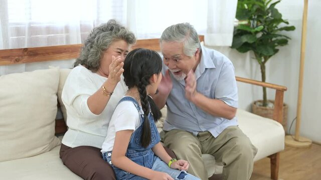 Asian elder couple play hide and seek with their granddaughter doing her homework with happily face in living room. Elder lifestyle, happy family, life insurance concept.