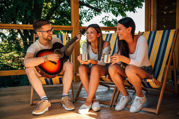 Young man playing guitar and two young women laughing. Friends on a summer holiday at countryside.