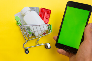 Online pharmacy. Closeup of hands on a phone and a mini shopping trolley with tablets. A woman chooses medicines on the Internet with home delivery