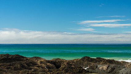 Fototapeta na wymiar Seascape with blue-green waves rising a beyond rocky foreground, and clouds on the horizon, beneath a blue sky. At Currumbin, Gold Coast, Queensland, Australia.