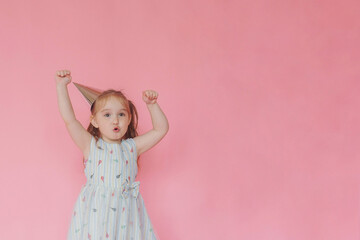 Obraz na płótnie Canvas Little cute happy girl in a birthday cap with a happy face rejoices raising his hands up on a pink background. Copy space