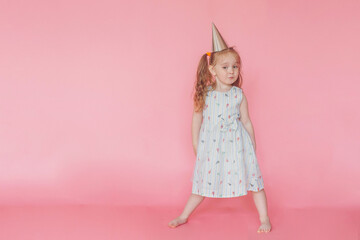 A little cute happy girl in birthday cap  grimaces with happy face on a pink background. Сhild birthday party. Copy space