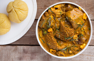 A white bowl of Nigerian dish Garri or eba and Oha soup cooked with assorted meat, beef, dried fish and cow belly meat 'shaki' decorated with green leaf spices and sliced yellow pepper on wood table