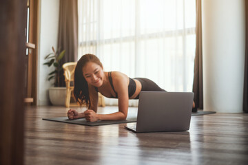 A beautiful young asian woman doing plank on training mat while watching online workout tutorials...