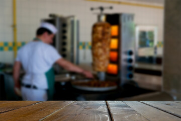 Wooden top of the table, desk, in the foreground.  Blurred image of a cook who fries meat for a shawarma in the background.